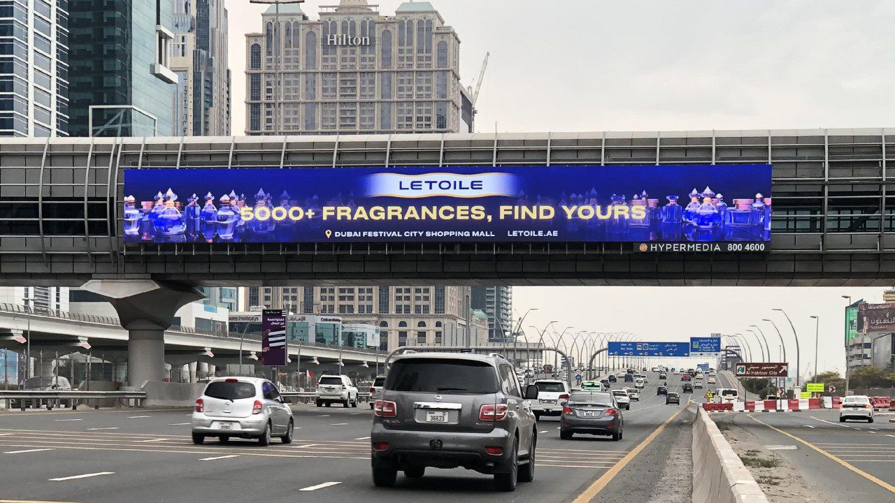 Large LED bridge banner advertising display on a major highway in Business Bay, Dubai, promoting a fragrance brand.